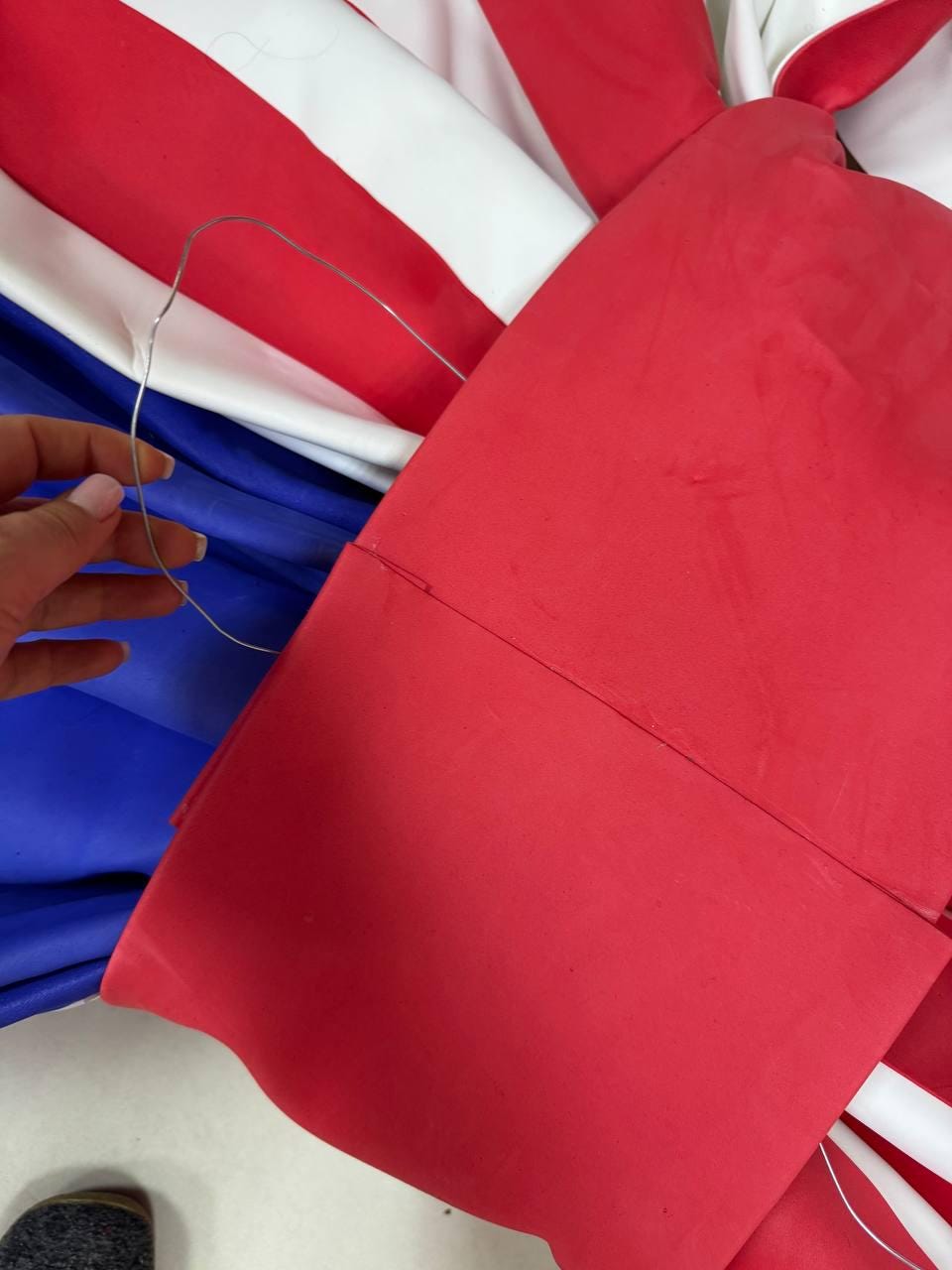 May include: A red, white, and blue fabric item, possibly a decorative piece or prop, with a wavy edge. The item is partially folded, revealing a blue section. A thin, silver wire is held near the fabric.