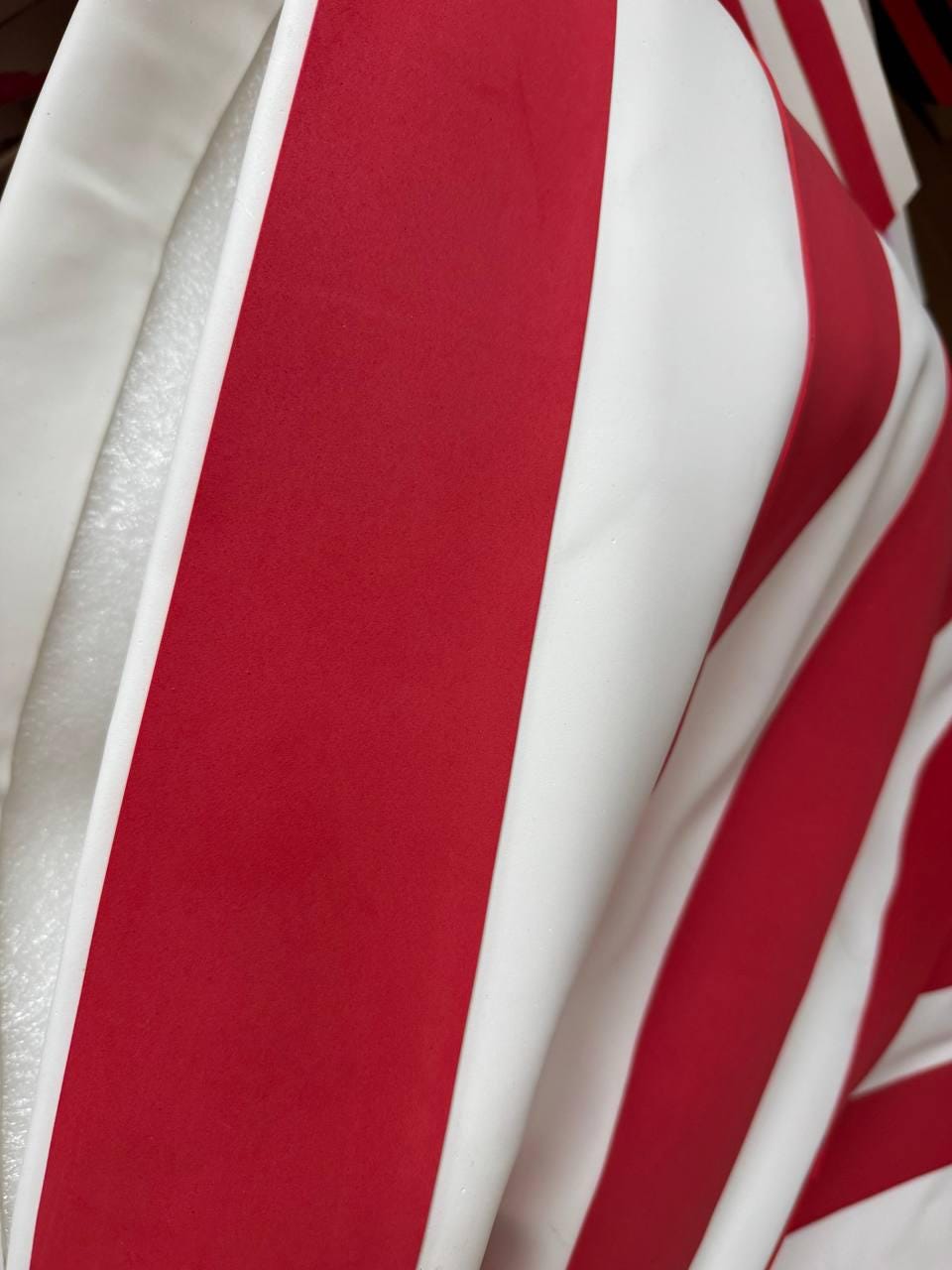 May include: Close-up of a fabric with bold red and white vertical stripes. The fabric appears to be smooth and possibly part of a garment or decorative item. The stripes are wide and evenly spaced.