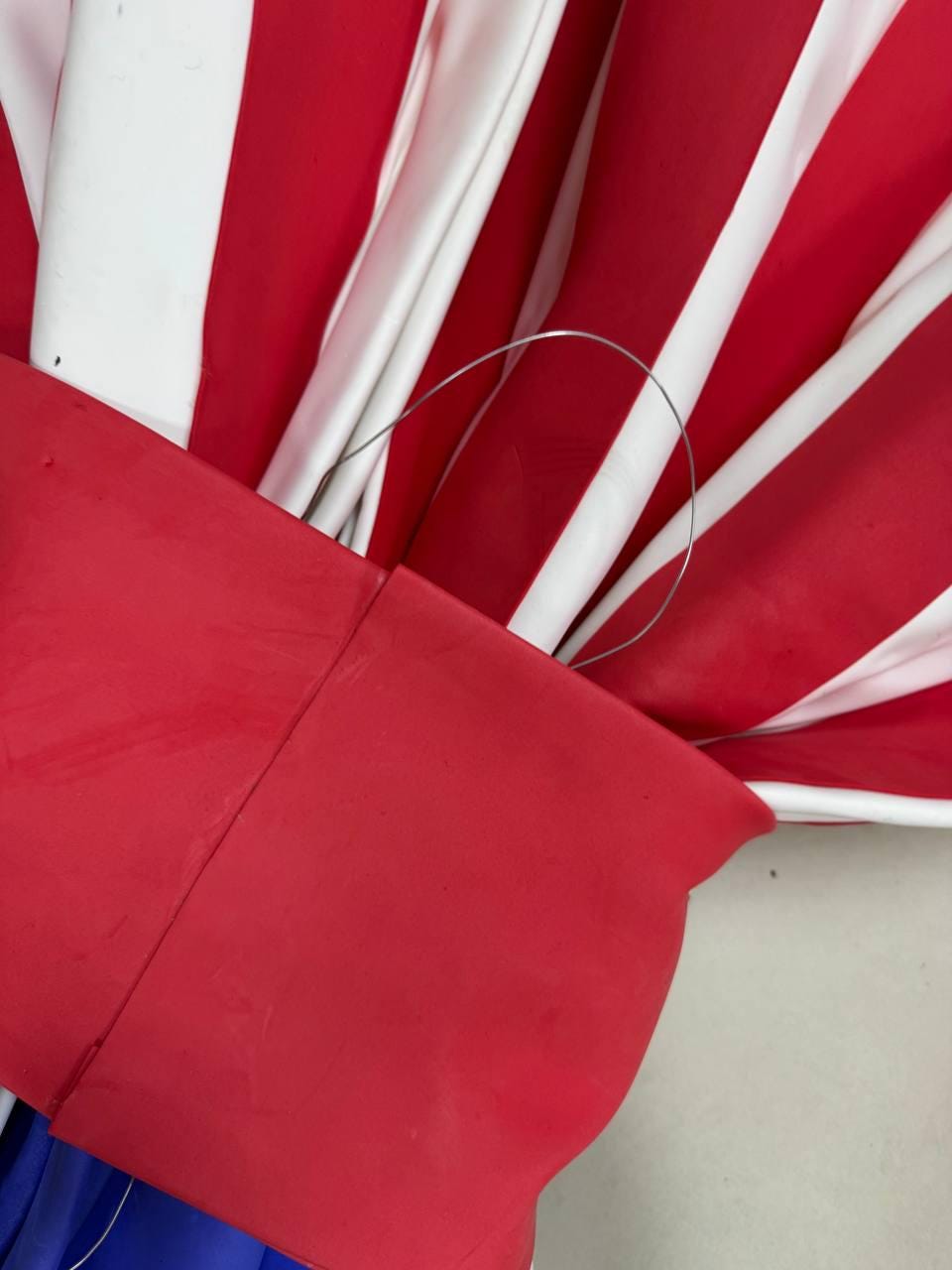 May include: Close-up of a red, white, and blue decorative item. The item features alternating red and white stripes, with a solid red section at the base. A silver wire handle is attached.