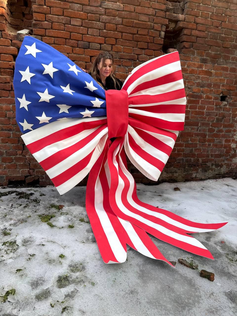May include: A large decorative bow in the style of the American flag. The bow features a blue section with white stars and red and white stripes. The bow is held in front of a brick wall.