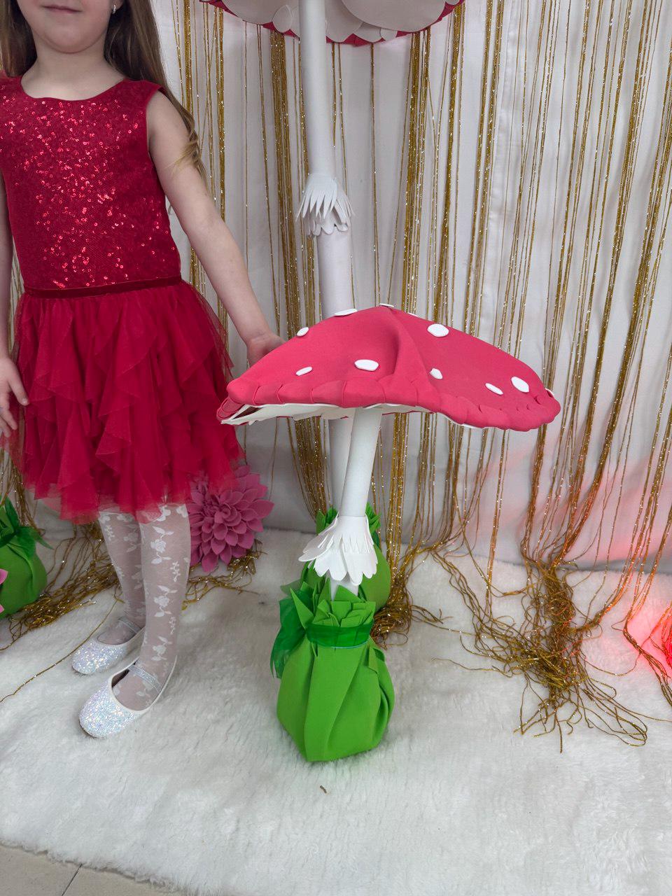 May include: A whimsical, oversized mushroom prop with a bright red cap and white polka dots. The mushroom has a white stem and is set on a green base. The background features a gold curtain.
