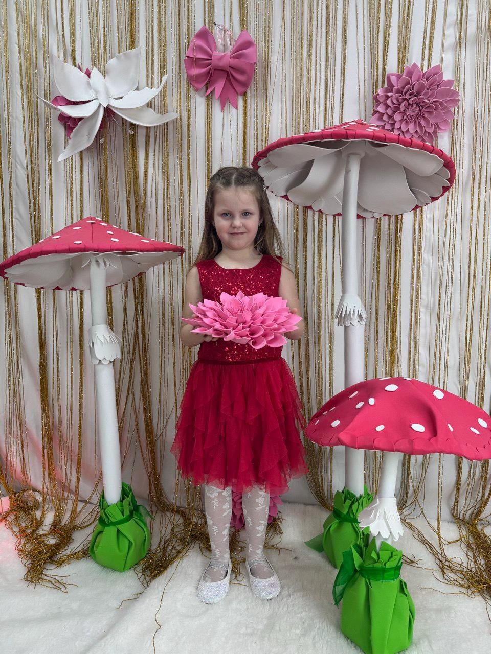 May include: A young girl in a red dress stands among whimsical mushroom decorations. The mushrooms have red caps with white spots and white stems. Pink flower and bow decorations adorn the background. The girl holds a large pink flower.