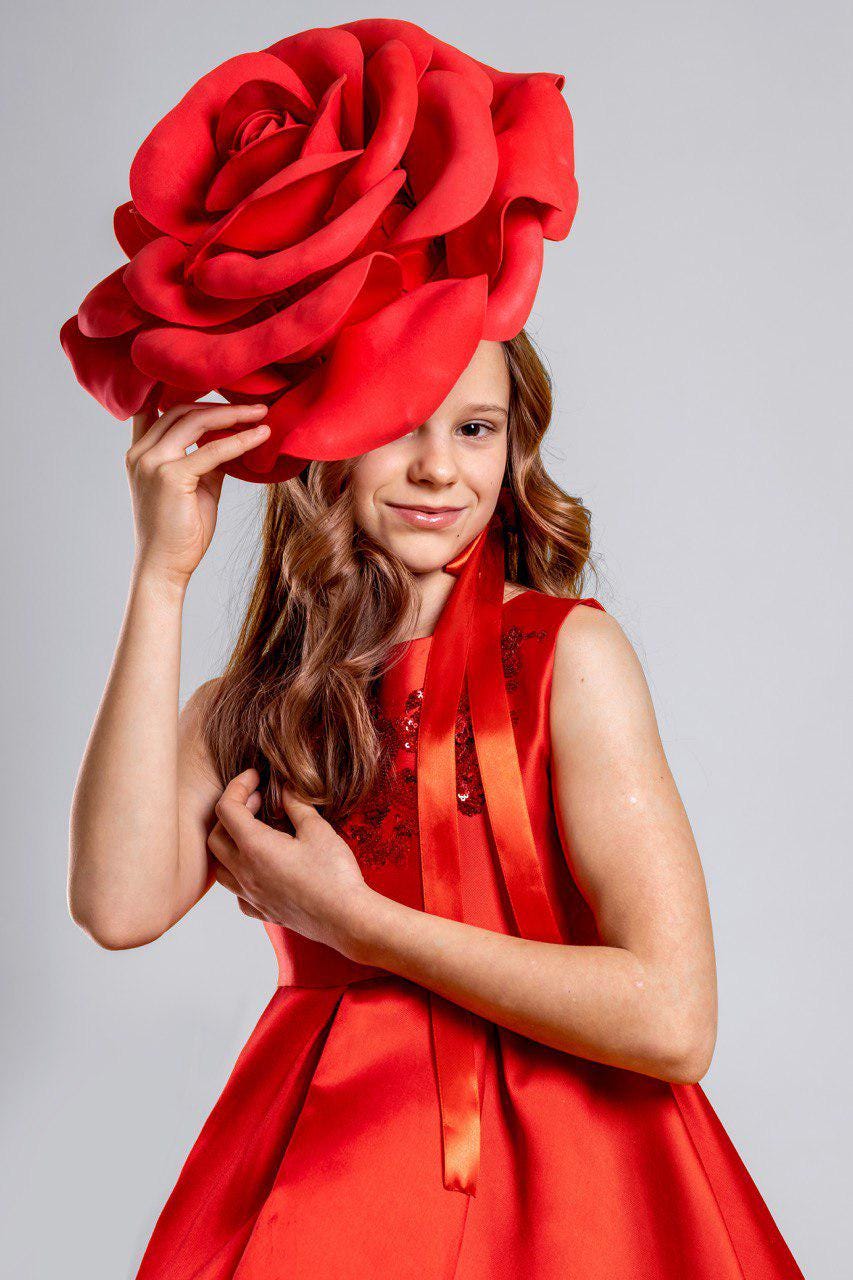 May include: A young person wearing a vibrant red dress and a large, oversized red rose headpiece. The dress features a fitted bodice and a flowing skirt. The headpiece is the focal point, creating a dramatic and eye-catching look.