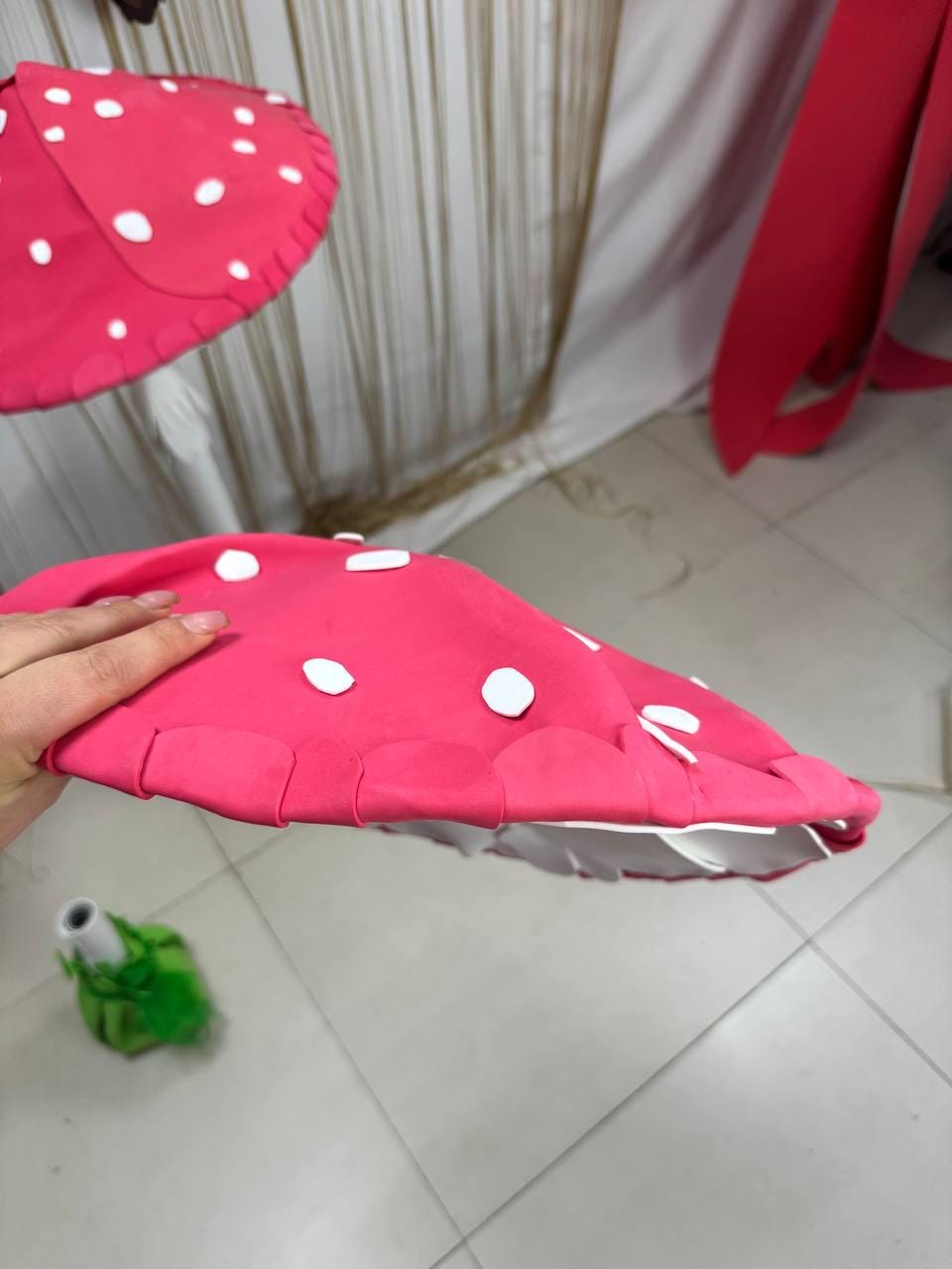 May include: A bright pink mushroom-shaped prop with white polka dots. The underside is white. The prop is held by a hand. A green and white object is in the background. The prop is likely for a costume or decoration.