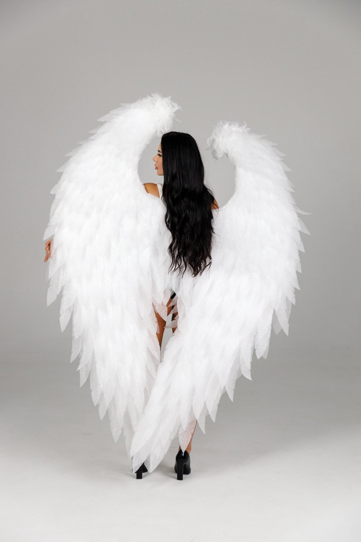 May include: A person with long black hair is seen from the back, wearing large white feathered angel wings. The wings are the focal point, with a detailed, layered design. The background is a plain, light gray. The person is wearing black heels.