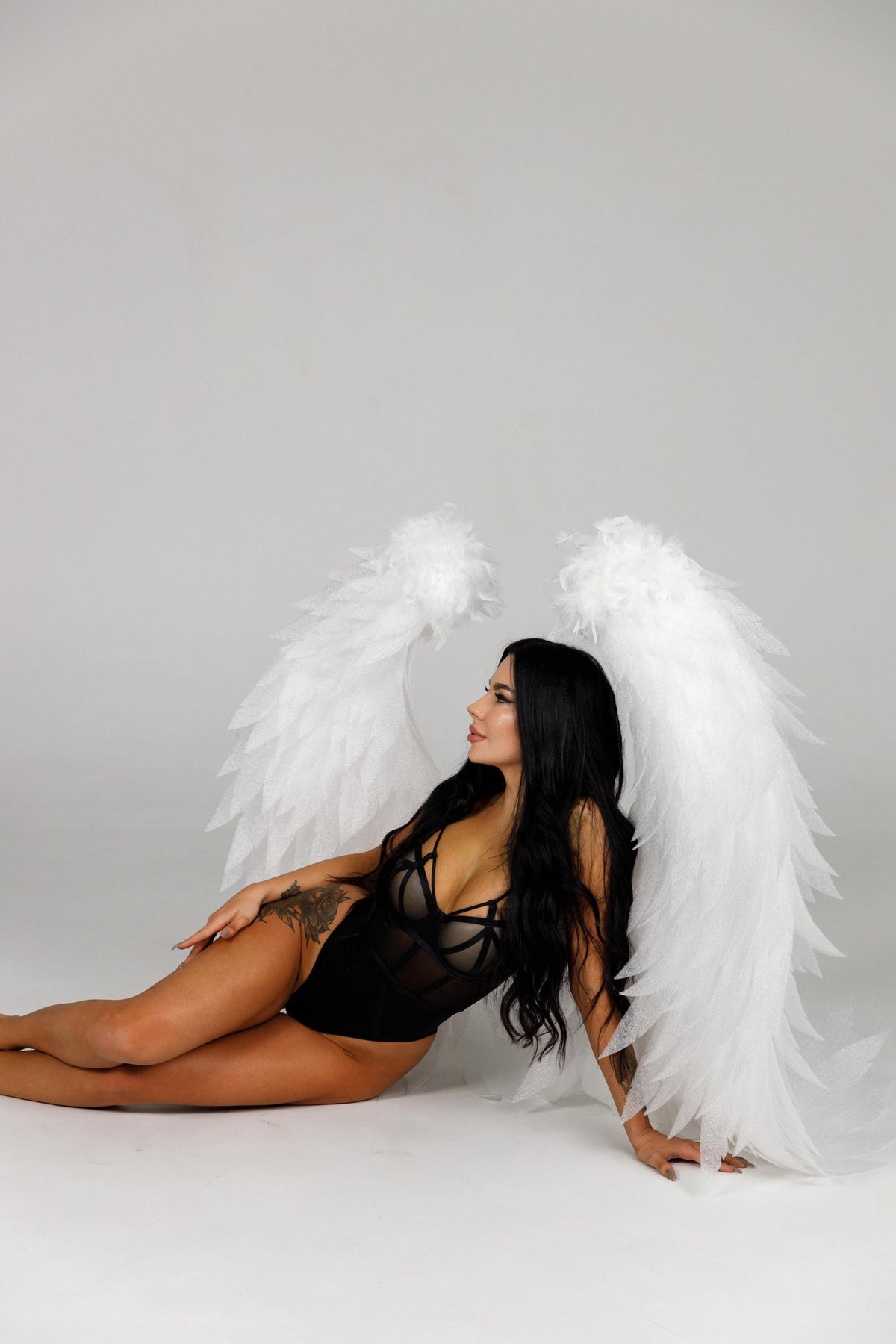 May include: A black bodysuit with a sheer panel and a strappy design. Large white feathered angel wings are behind the bodysuit. The model has long black hair and is lying down on a white surface.