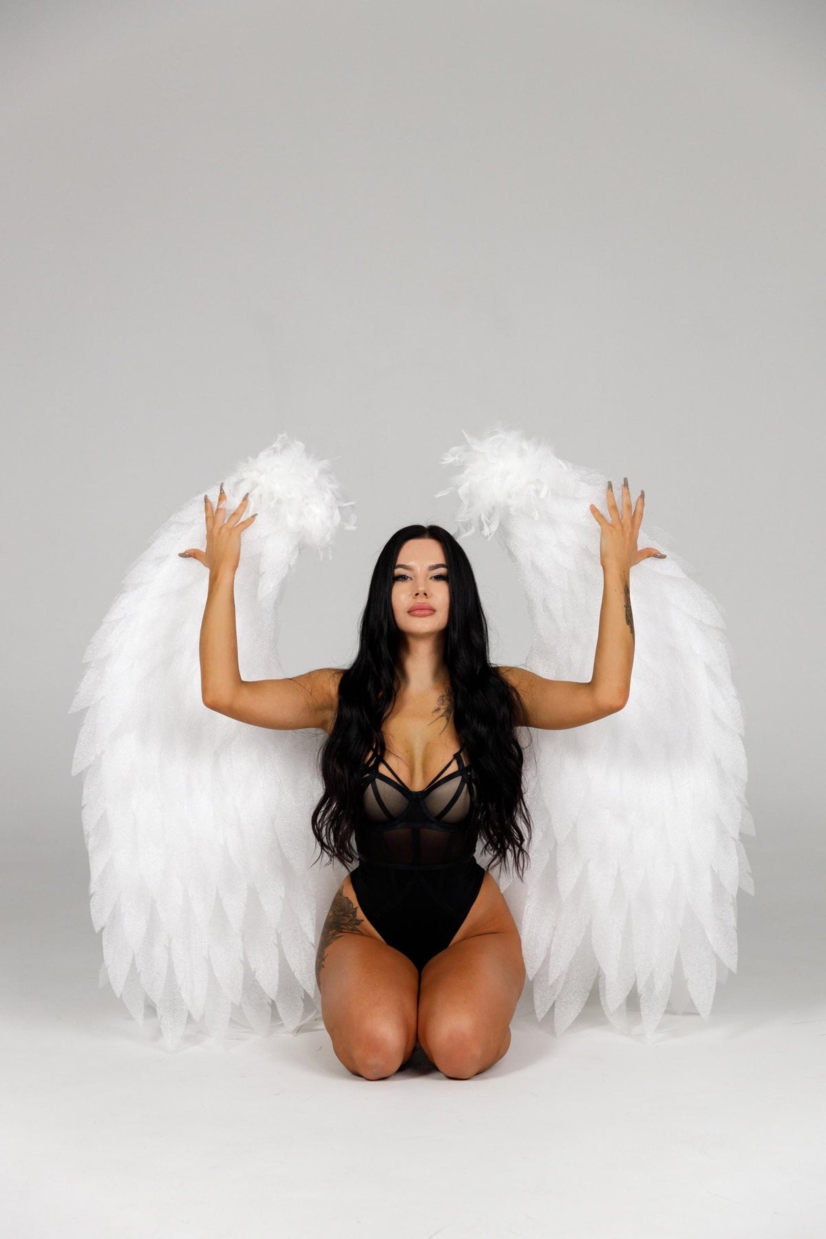 May include: A person kneels, wearing a black bodysuit and large white feathered angel wings. The person has long black hair and is posing with arms raised. The background is a plain white surface.
