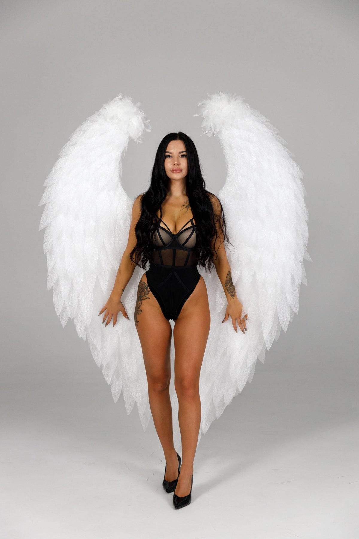 May include: A person wearing a black bodysuit with a sheer panel and black high heels, posing in front of large white feathered angel wings. The wings are symmetrical and spread wide, creating a dramatic effect against a gray backdrop.