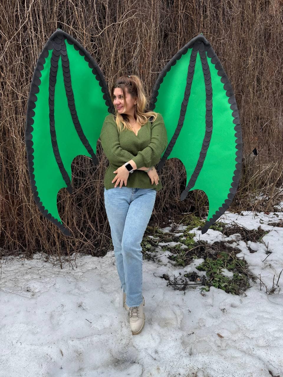 May include: Large green dragon wings with black trim. The wings are worn by a person wearing a green sweater and blue jeans. The wings are made of a lightweight material and are designed to be worn on the back.