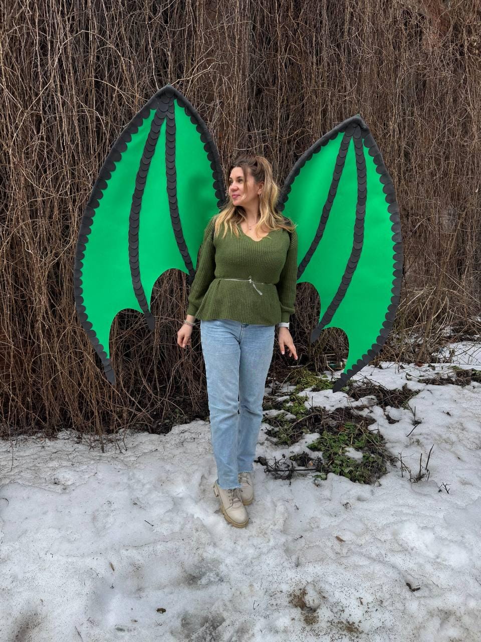 May include: Large green dragon wings with black trim. The wings are made of a lightweight material and are designed to be worn. The wings are a vibrant green color and have a detailed, textured appearance. The wings are set against a backdrop of brown branches and snow.