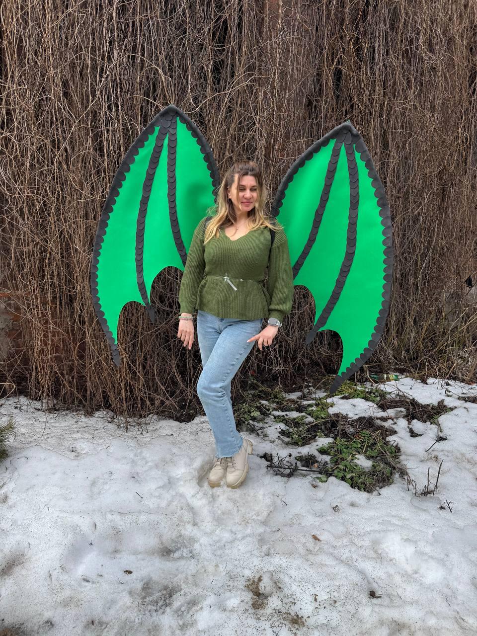 May include: A person wearing large, green dragon wings with black trim. The wings are made of a sturdy material and are designed to be worn on the back. The person is wearing a green sweater and blue jeans, standing in a snowy outdoor setting.