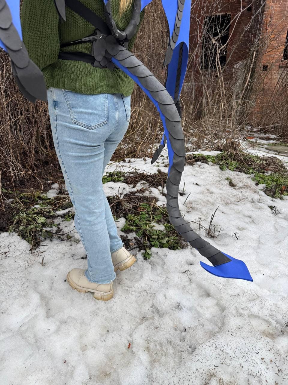 May include: A costume piece featuring a black and gray dragon tail with blue accents. The tail is attached to a harness. The costume is worn outdoors in a snowy environment.
