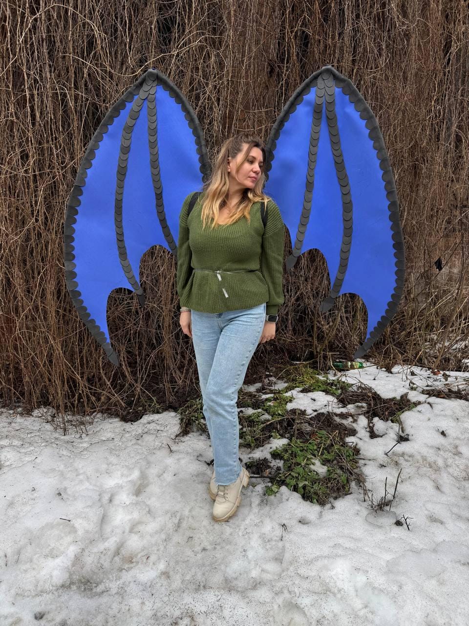 May include: A person wearing a green sweater and light blue jeans stands in front of large blue dragon wings with black trim. The wings are spread out, creating a fantasy or cosplay theme. The background includes bare branches and snow.
