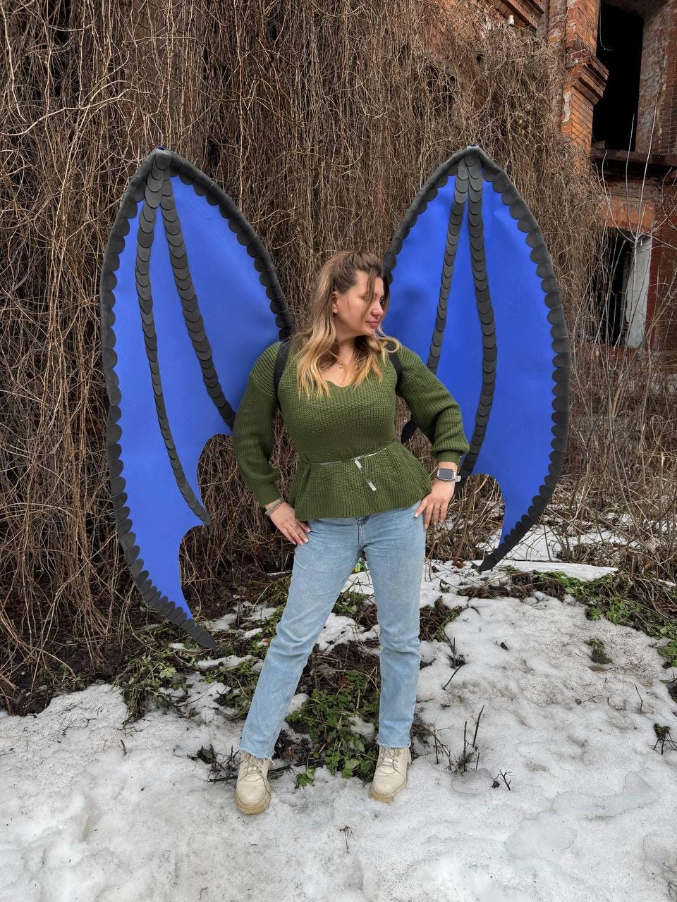 May include: Blue and black dragon wings with a scalloped edge. The wings are worn by a person wearing a green sweater and light blue jeans. The wings are a costume accessory.