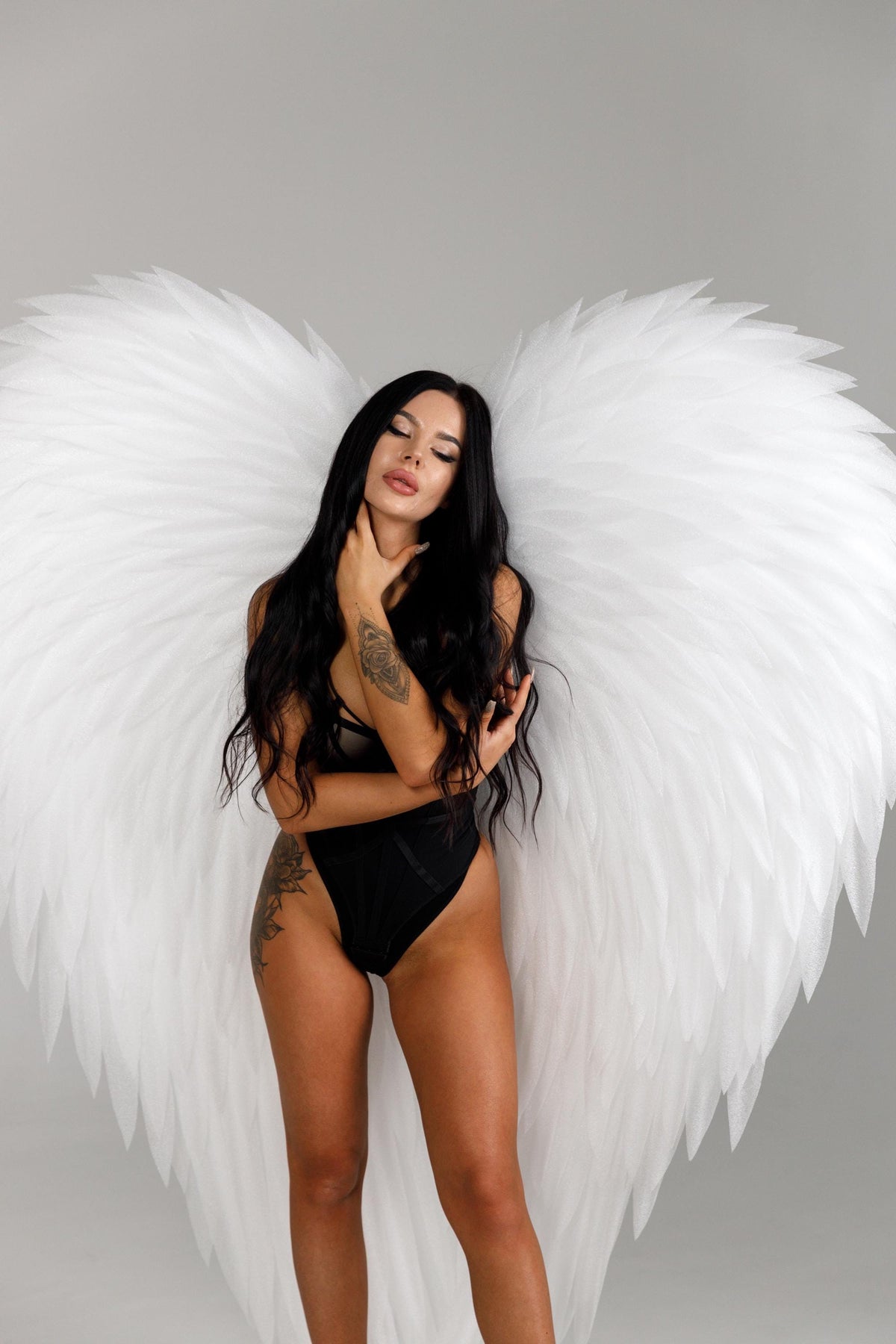 May include: A person wearing a black bodysuit and large white angel wings. The wings are feathered and spread wide. The person has long black hair and tattoos on their arms and legs.