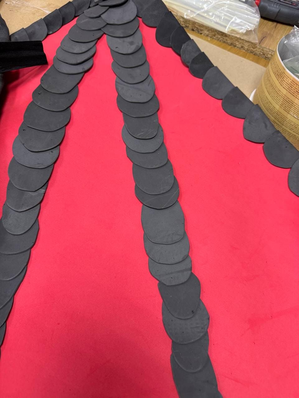 May include: A close-up view of a craft project in progress. The image shows a vibrant red surface with rows of dark gray, circular, scale-like shapes arranged in a fan-like pattern. The scales are made of a flexible material.