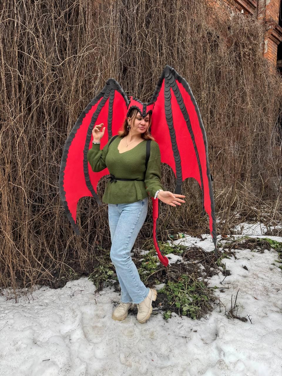 May include: A costume featuring large red and black dragon wings, a matching headpiece, and a red tail. The wings are spread wide, and the costume is worn outdoors. The person is wearing a green sweater and blue jeans.