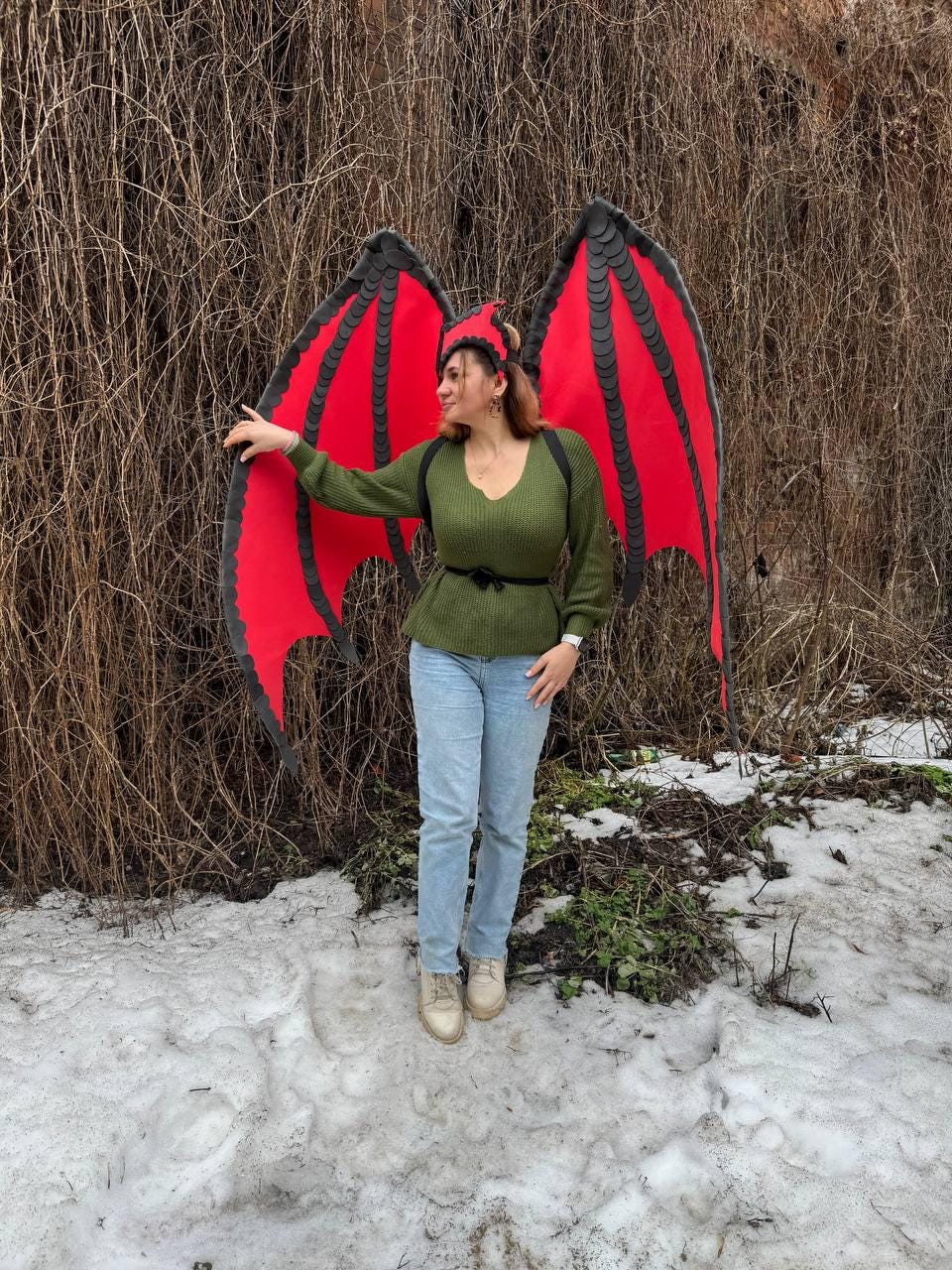 May include: A person wearing a green sweater, blue jeans, and beige shoes stands outdoors, posing with large red and black dragon wings. A matching red and black headpiece completes the costume. The background features bare branches and snow.