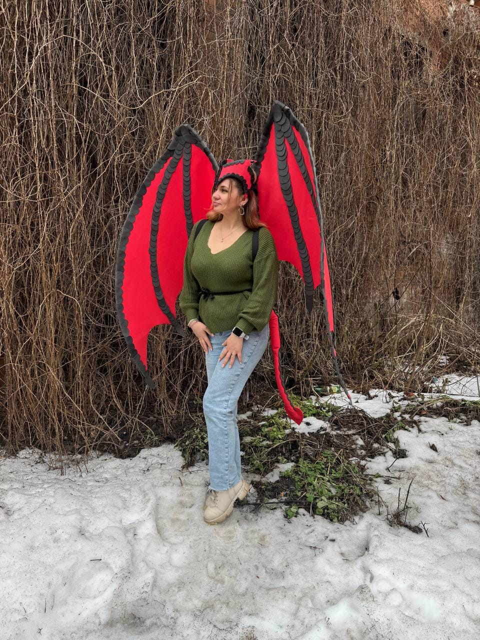 May include: A person wearing a red and black dragon wing costume with a matching headpiece and tail. The wings are large and detailed, with a red membrane and black edging. The costume is worn with a green sweater and blue jeans.