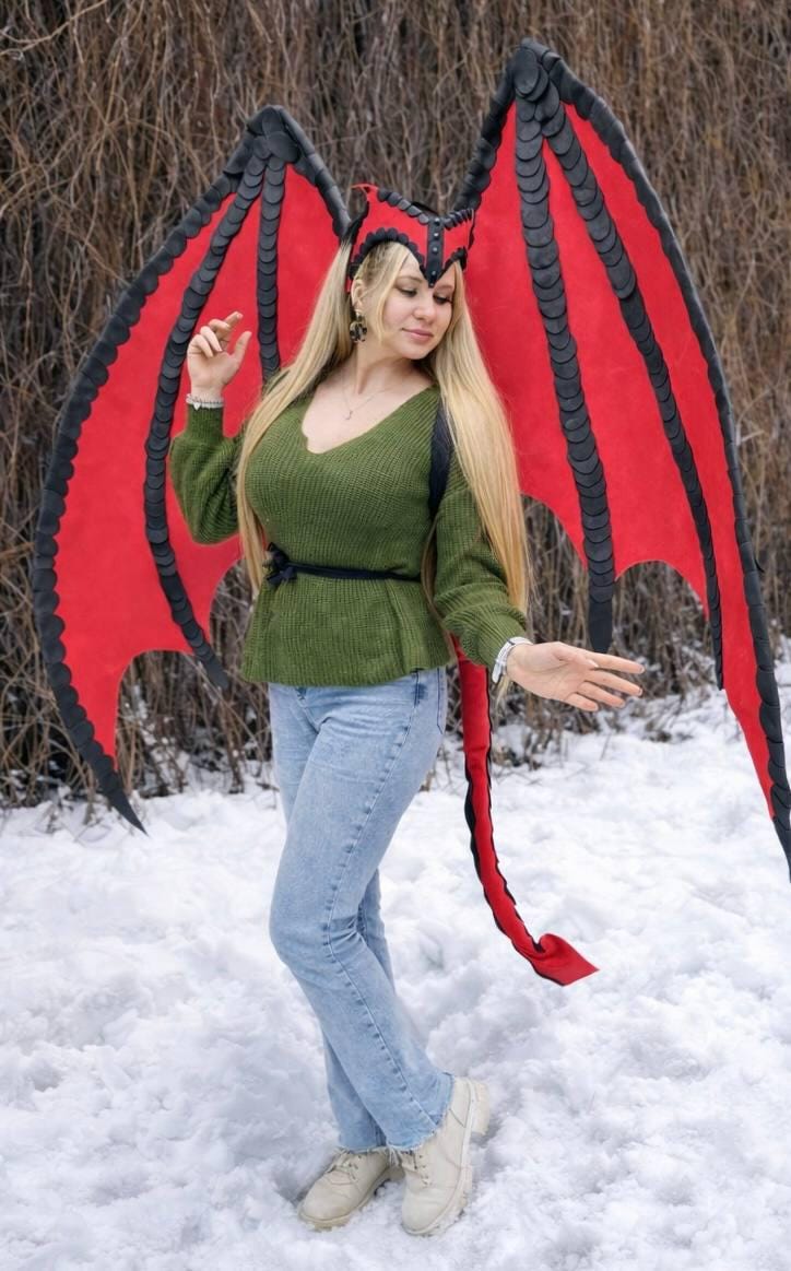 May include: A person wearing a green sweater and blue jeans, posing outdoors in the snow. The person is wearing a red and black dragon wing costume with a matching headpiece and tail. The wings are large and detailed.