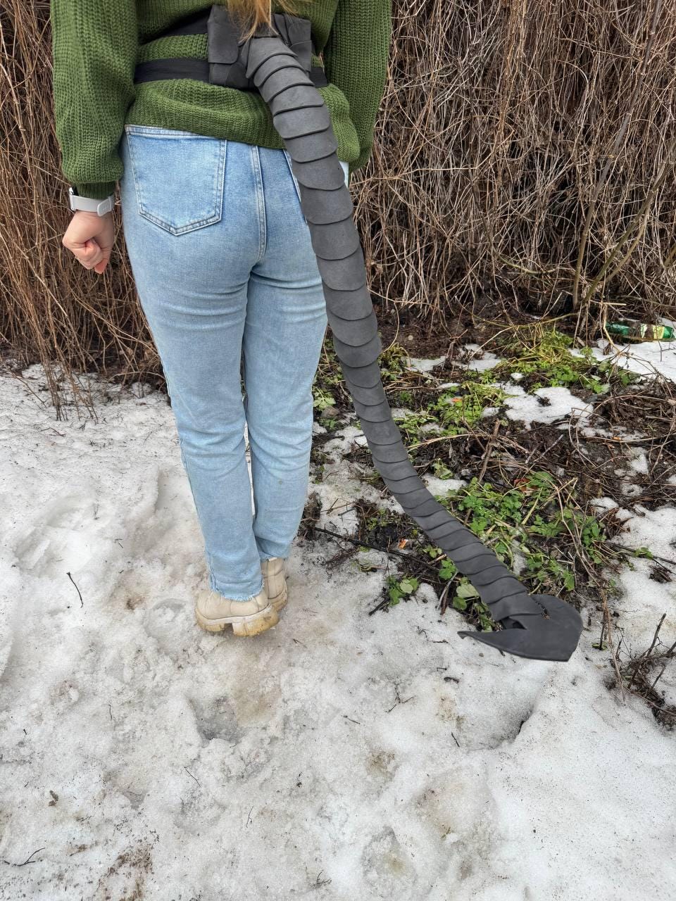 May include: A gray, segmented costume tail attached to a black belt. The tail is long and flexible, with a pointed tip. The person is wearing a green sweater, light blue jeans, and beige boots, standing in a snowy area.