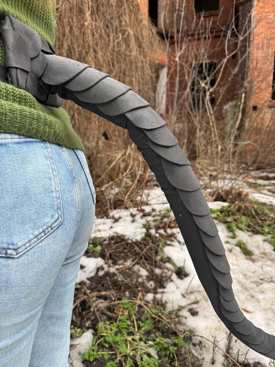May include: A black dragon tail prop with scale-like details. The tail is curved and flexible, with a dark color scheme. The prop is designed to be worn, with a portion of the tail attached to a person wearing a green sweater and blue jeans.