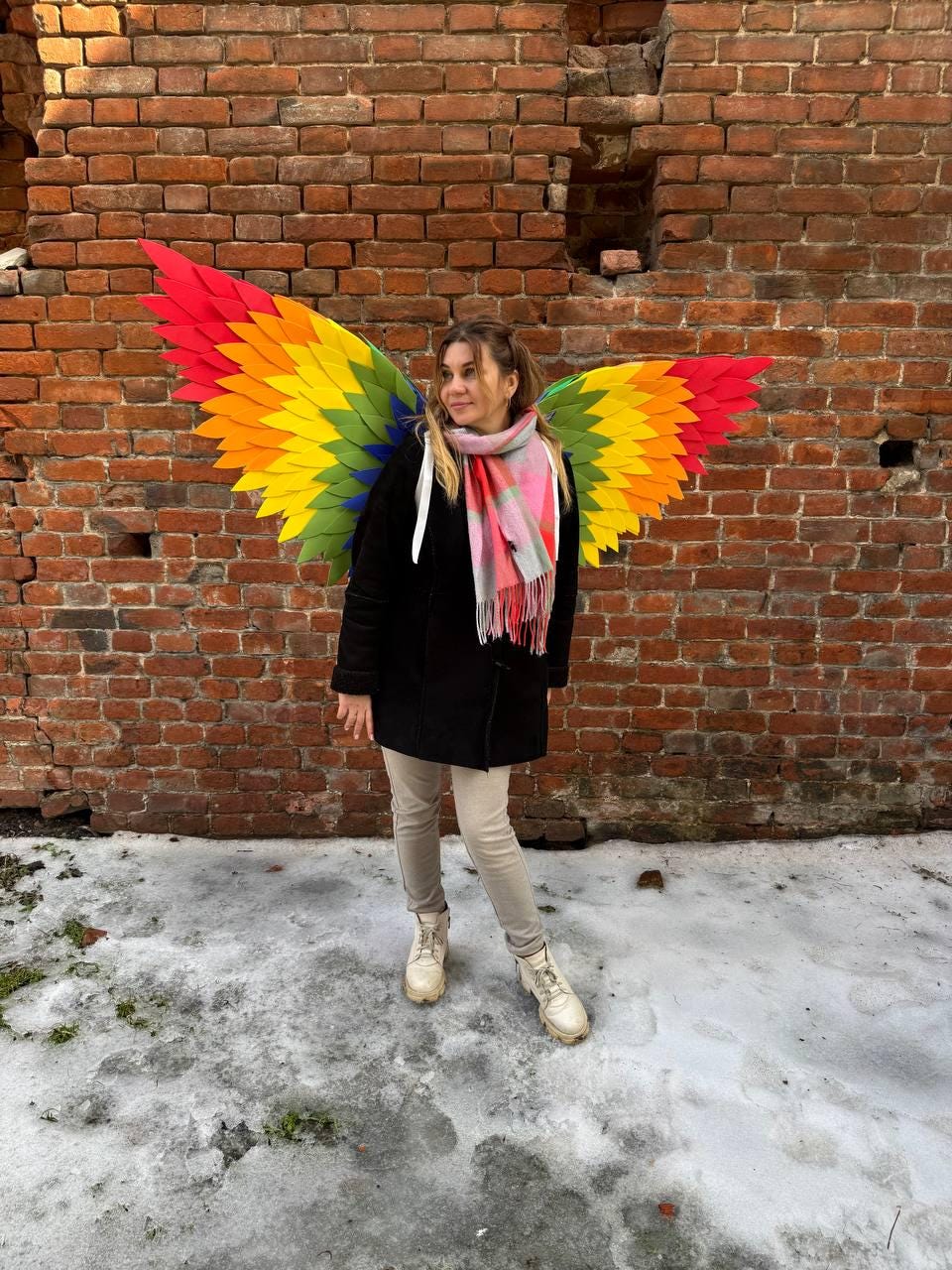 May include: A person wearing colorful rainbow-colored wings, standing in front of a brick wall. The wings feature a vibrant gradient of red, orange, yellow, green, and blue. The person is wearing a black coat, light-colored pants, and white boots.