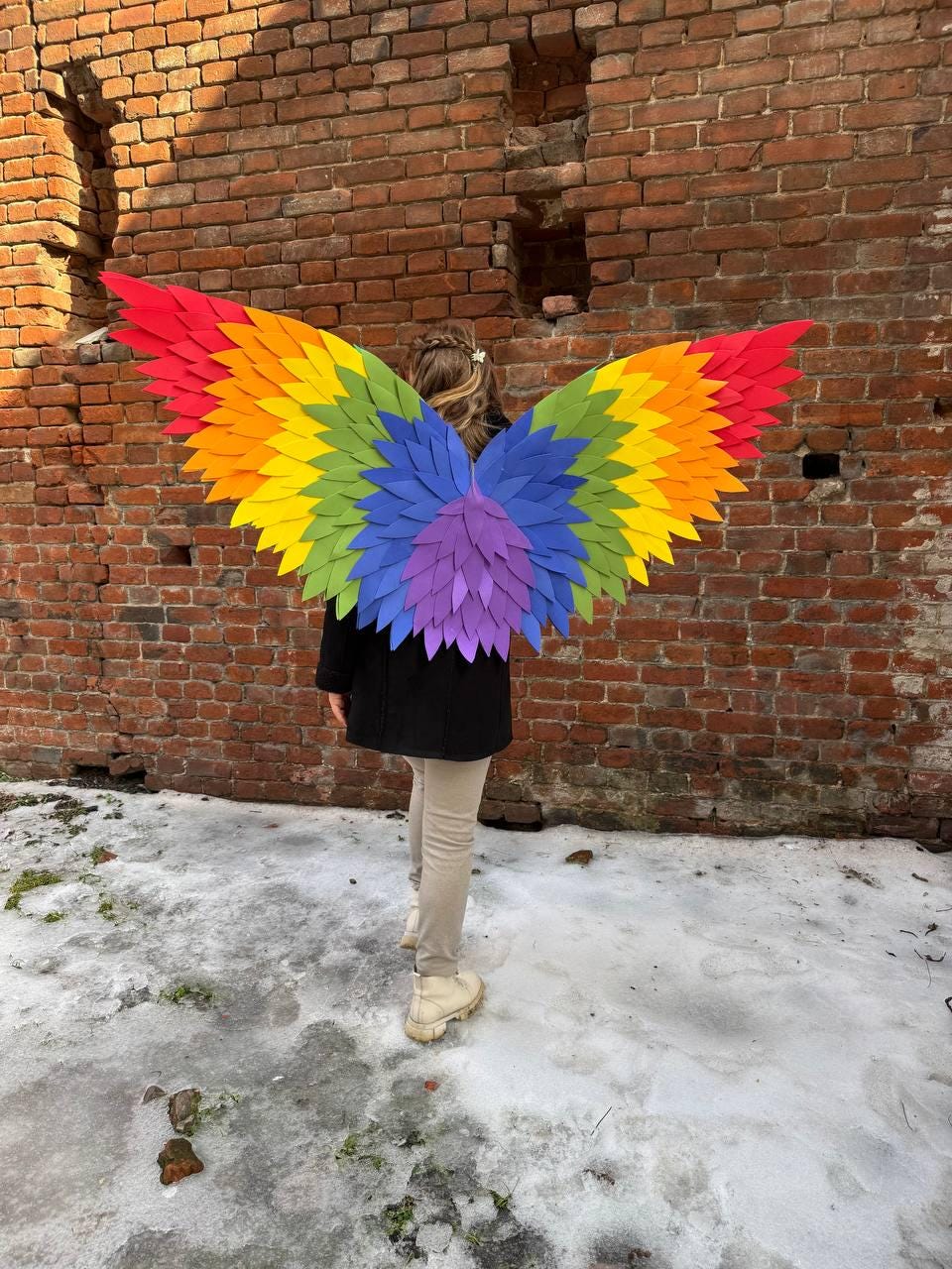 May include: A person wearing colorful rainbow-colored wings. The wings are made of layered paper in red, orange, yellow, green, blue, and purple. The person is standing in front of a brick wall.