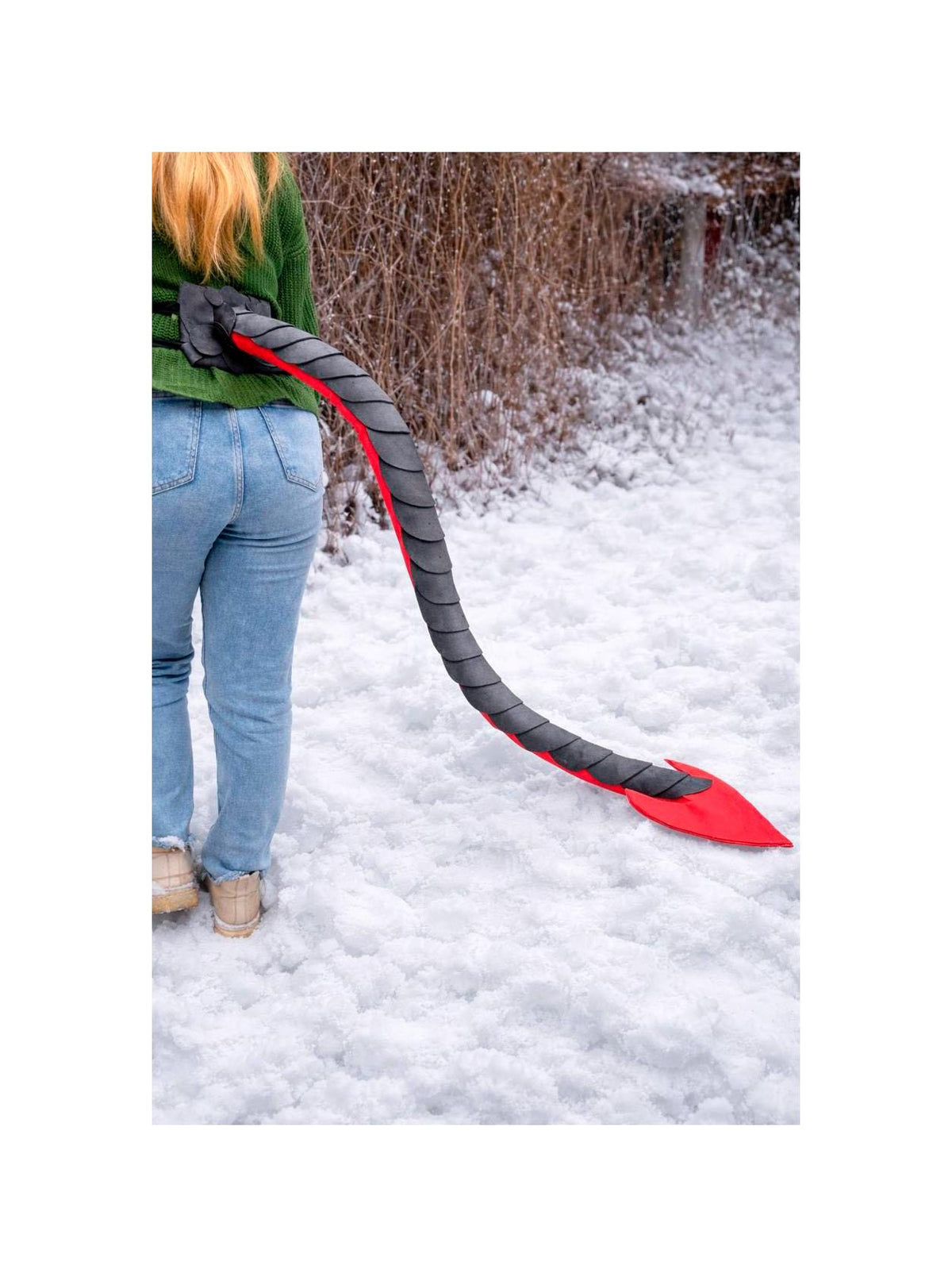 May include: A gray and red devil tail prop, worn around the waist, extends down towards the snow. The tail is gray with red accents and a red tip. The person is wearing a green sweater and blue jeans.