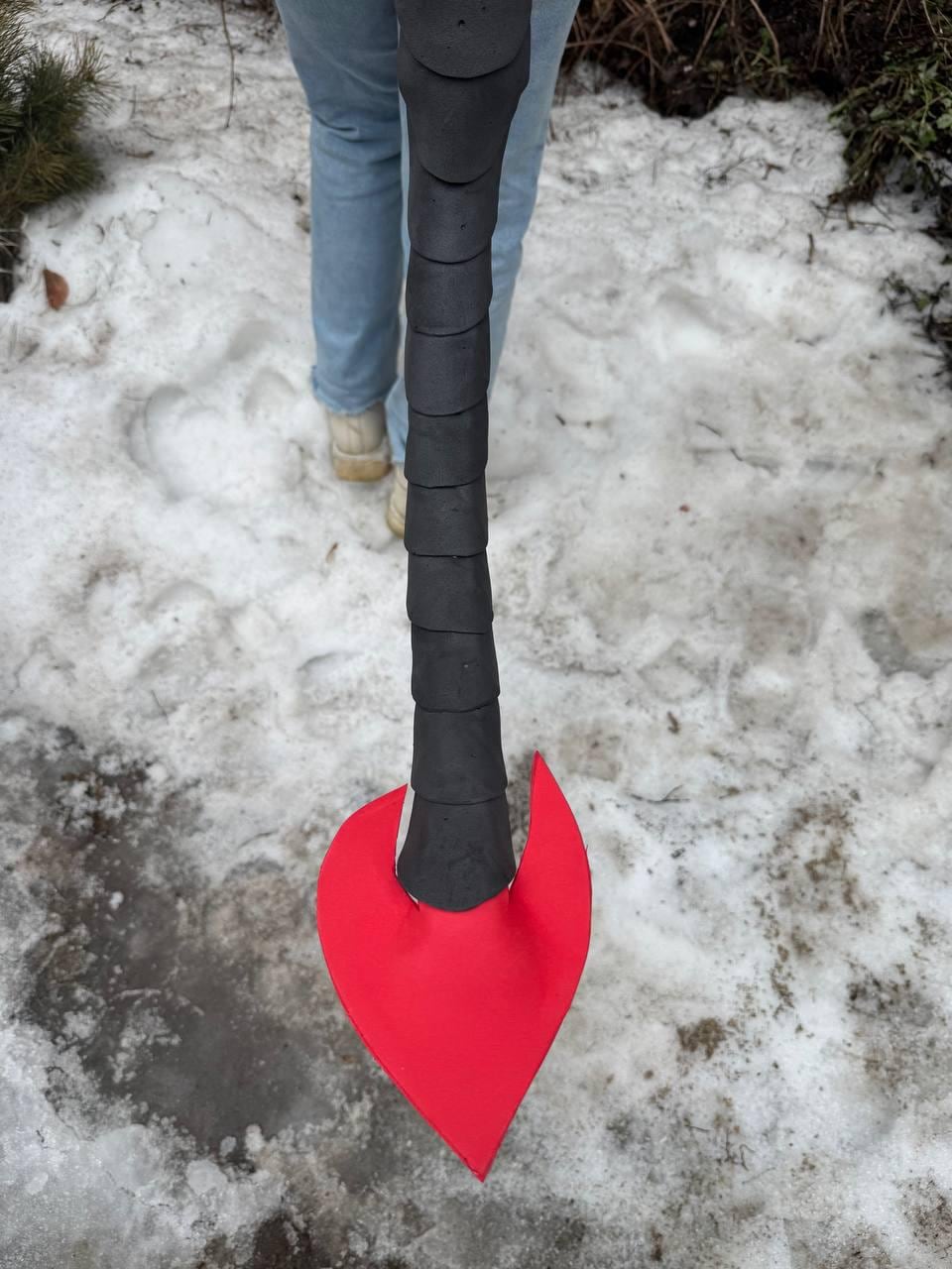 May include: A black and red prop weapon, possibly a fantasy axe or spear, held by a person. The weapon has a black handle with a textured design and a bright red, heart-shaped blade. The background shows snow and a person wearing blue jeans.