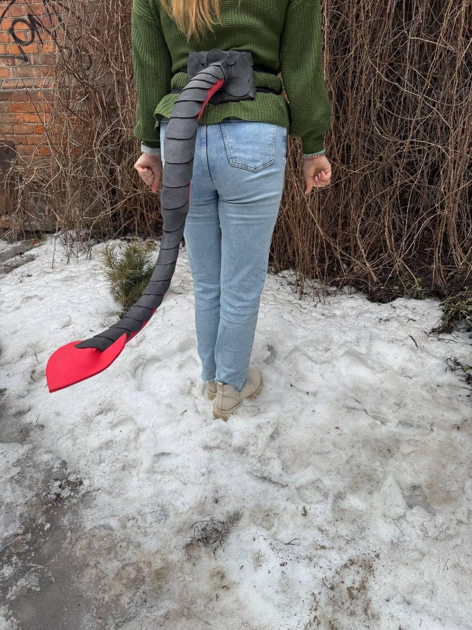 May include: A costume tail, featuring a segmented gray design with a red underside and a red, paddle-shaped tip. The tail is attached to a black belt. The person is wearing a green sweater and blue jeans.