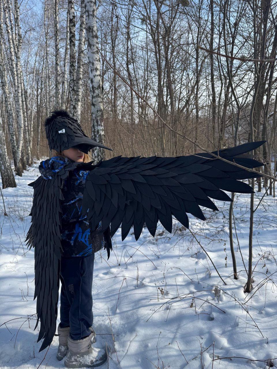 May include: A black raven costume with large, detailed wings and a matching hat with a beak. The costume is worn in a snowy forest setting, creating a striking contrast against the white snow and bare trees. The wings are spread wide.