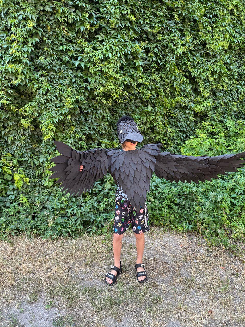 May include: A child wearing a black raven costume with large, outstretched wings. The costume includes a feathered headpiece and a body piece with layered, scale-like details. The child is wearing black sandals and patterned shorts.