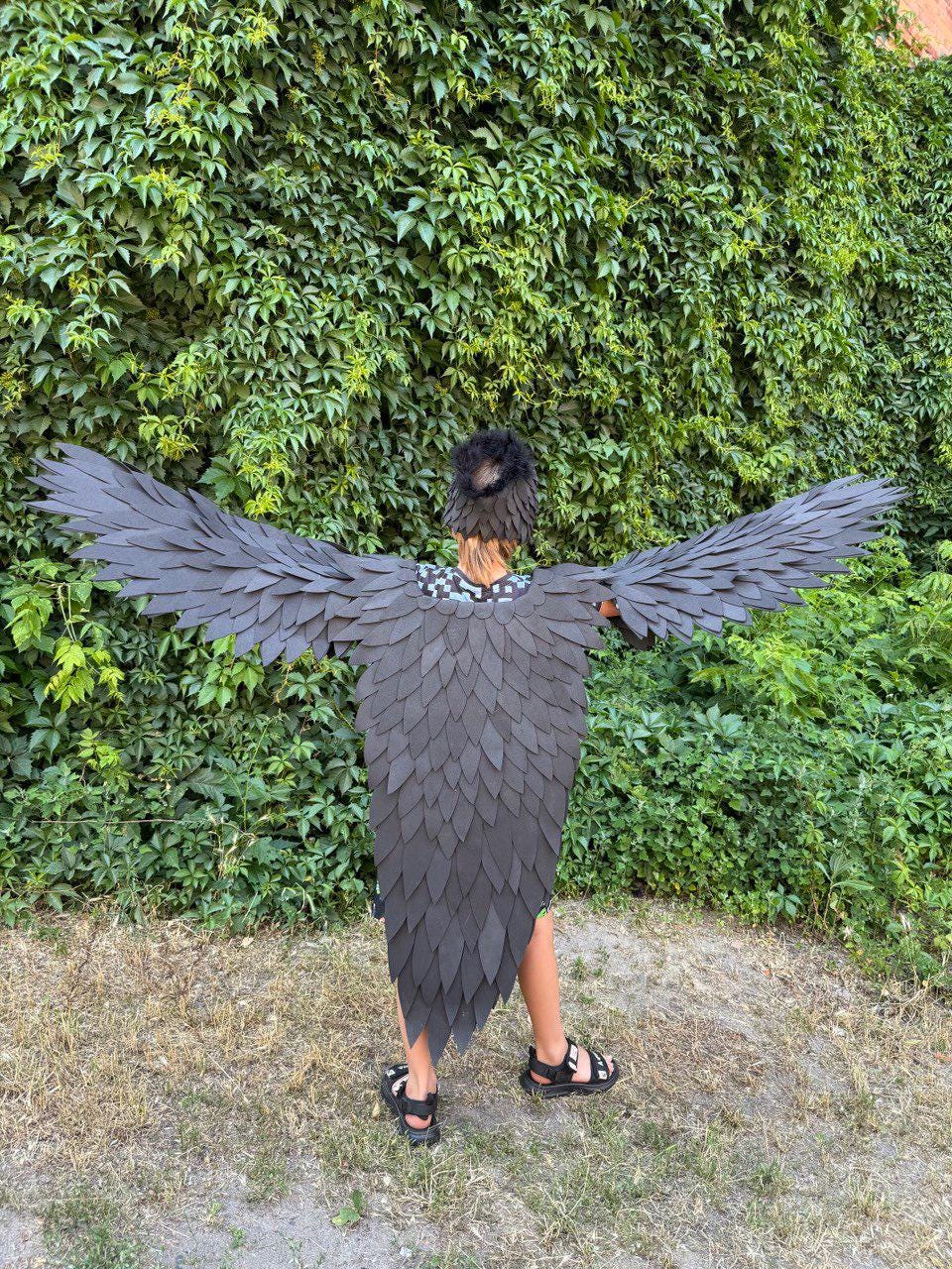 May include: A black raven costume with large, detailed wings and a matching headpiece. The costume features a layered design, creating a feathered effect. The wings are spread wide, and the costume is worn outdoors against a backdrop of green foliage.