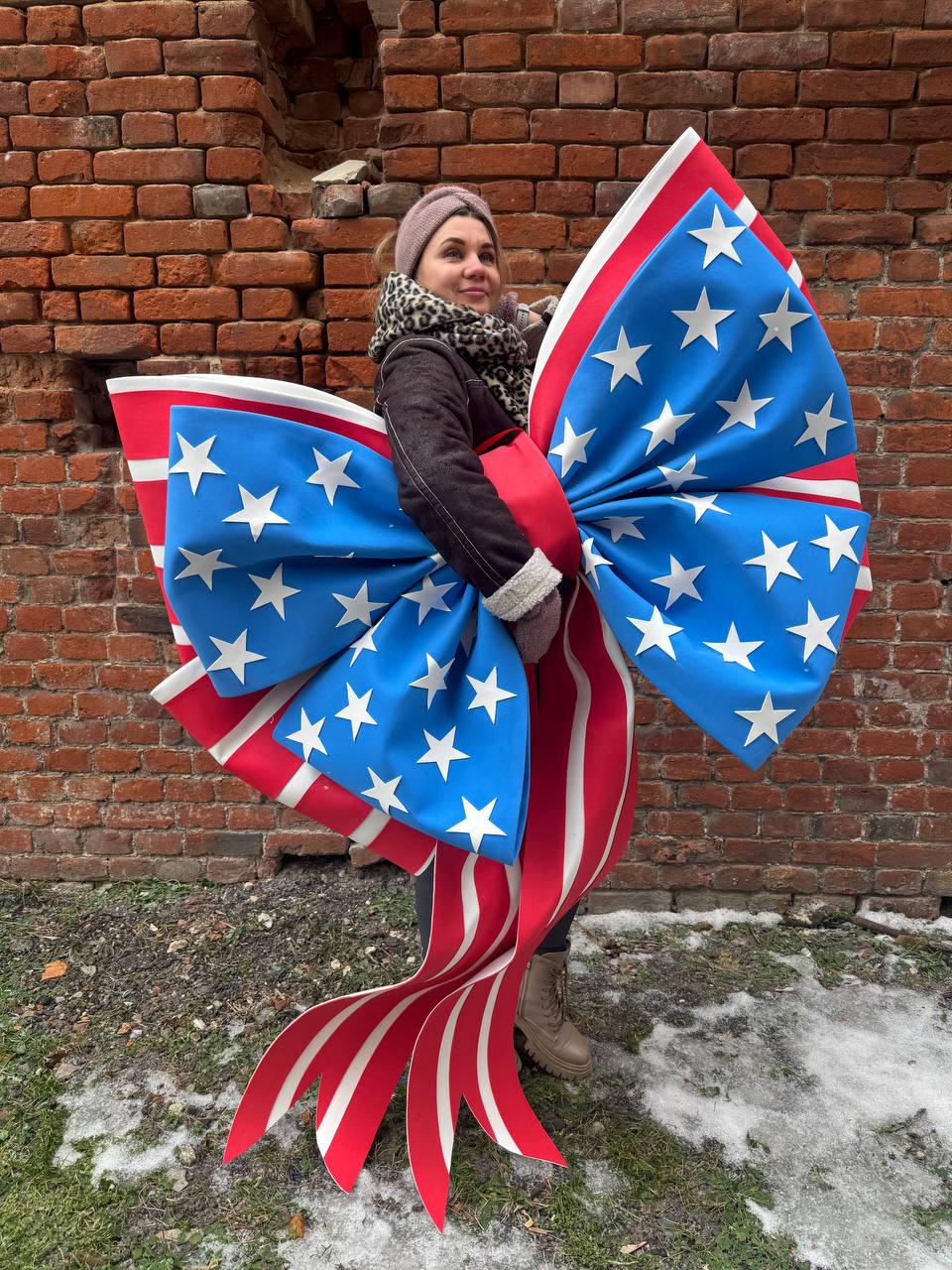 May include: A large decorative bow featuring the American flag design. The bow has a blue center with white stars and red and white striped ribbons. The bow is held in front of a brick wall.