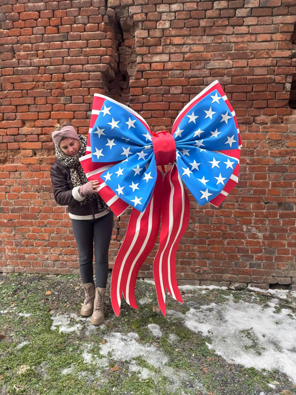 May include: A large decorative bow with a patriotic design. The bow features a blue center with white stars, red and white striped ribbons, and a red center knot. The bow is displayed in front of a brick wall.