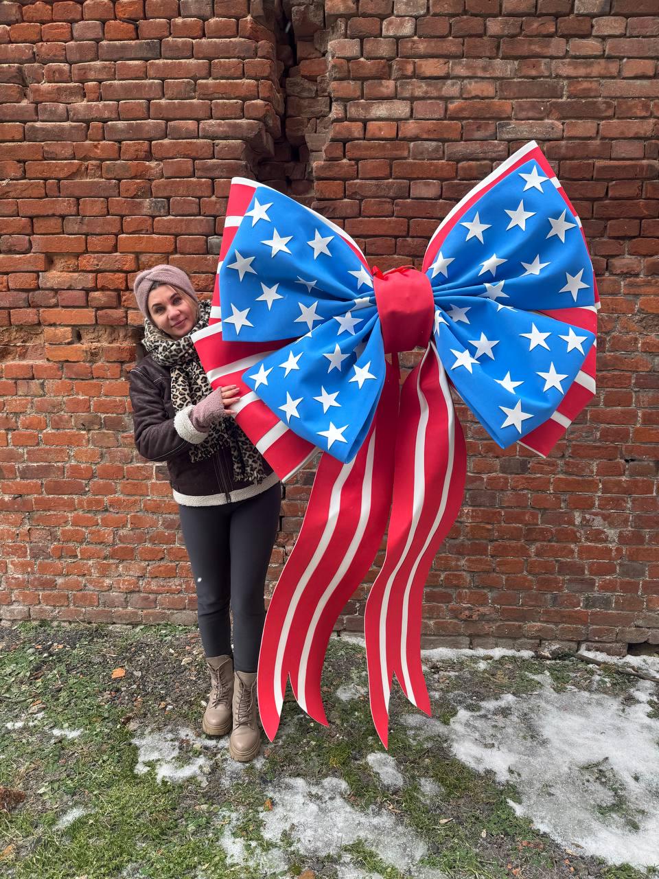 May include: A large decorative bow in the style of the American flag. The bow features a blue center with white stars, red and white striped edges, and long red and white striped ribbons. The bow is approximately 5 feet wide.