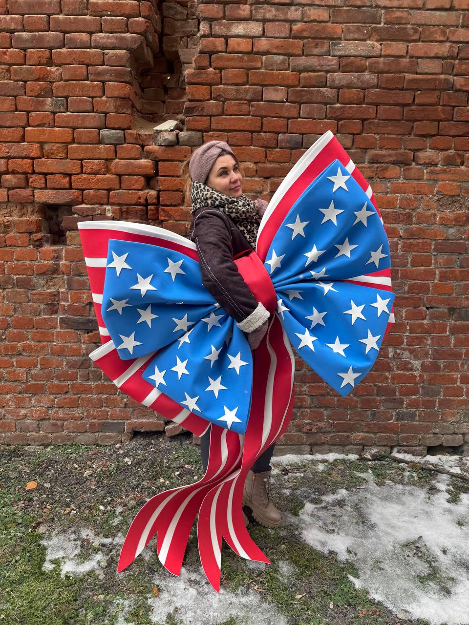 May include: A large decorative bow featuring a patriotic design with blue sections adorned with white stars, red and white striped ribbons. The bow is held in front of a brick wall.