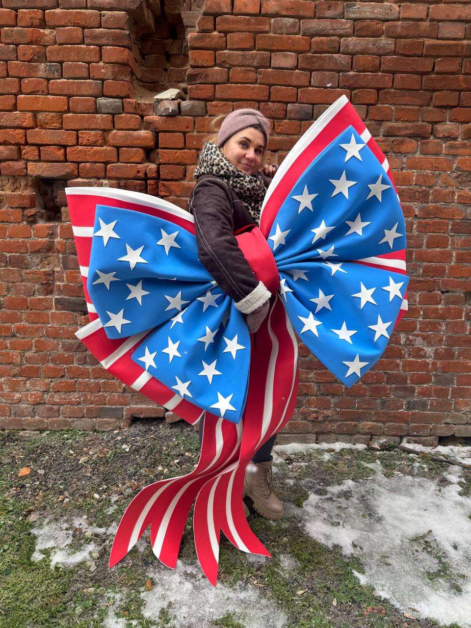 May include: A large decorative bow in the style of the American flag. The bow features a blue center with white stars, red and white striped ribbons, and a red center. The bow is held in front of a brick wall.