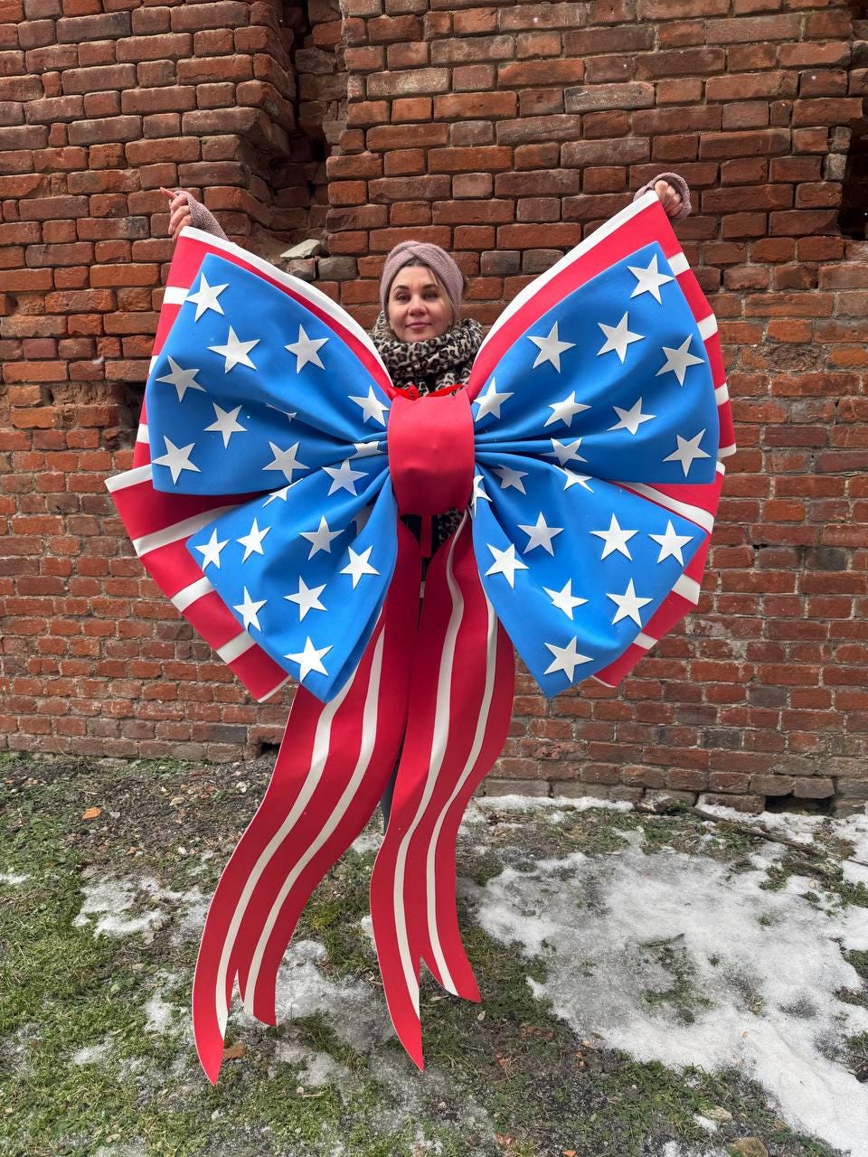 May include: A large decorative bow featuring the American flag design. The bow has a blue center with white stars, red and white striped ribbons, and a red center knot. The bow is held up against a brick wall.