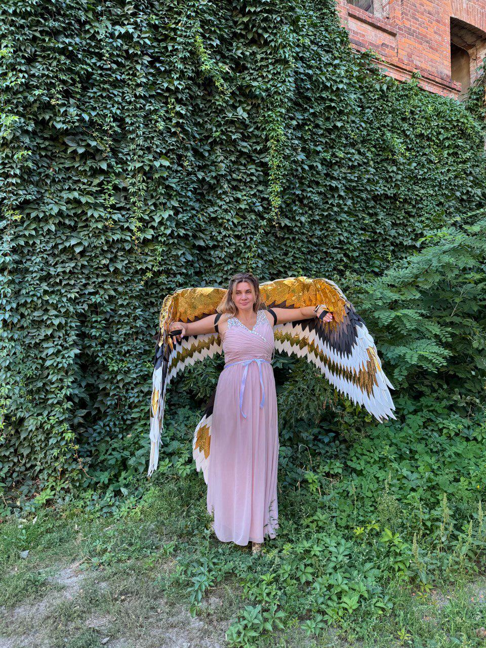 May include: A person wearing a long, light pink dress and large, decorative wings with gold, black, and white feathers. The wings are spread wide against a backdrop of lush green foliage and a brick building.