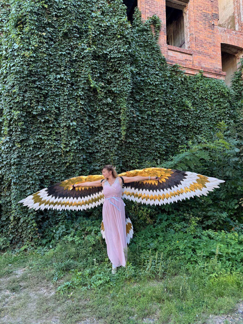 May include: A woman wearing a long, light pink dress and large, decorative wings with gold, brown, black, and white feathers. The wings are spread wide, and the woman is standing in front of a wall covered in green ivy.