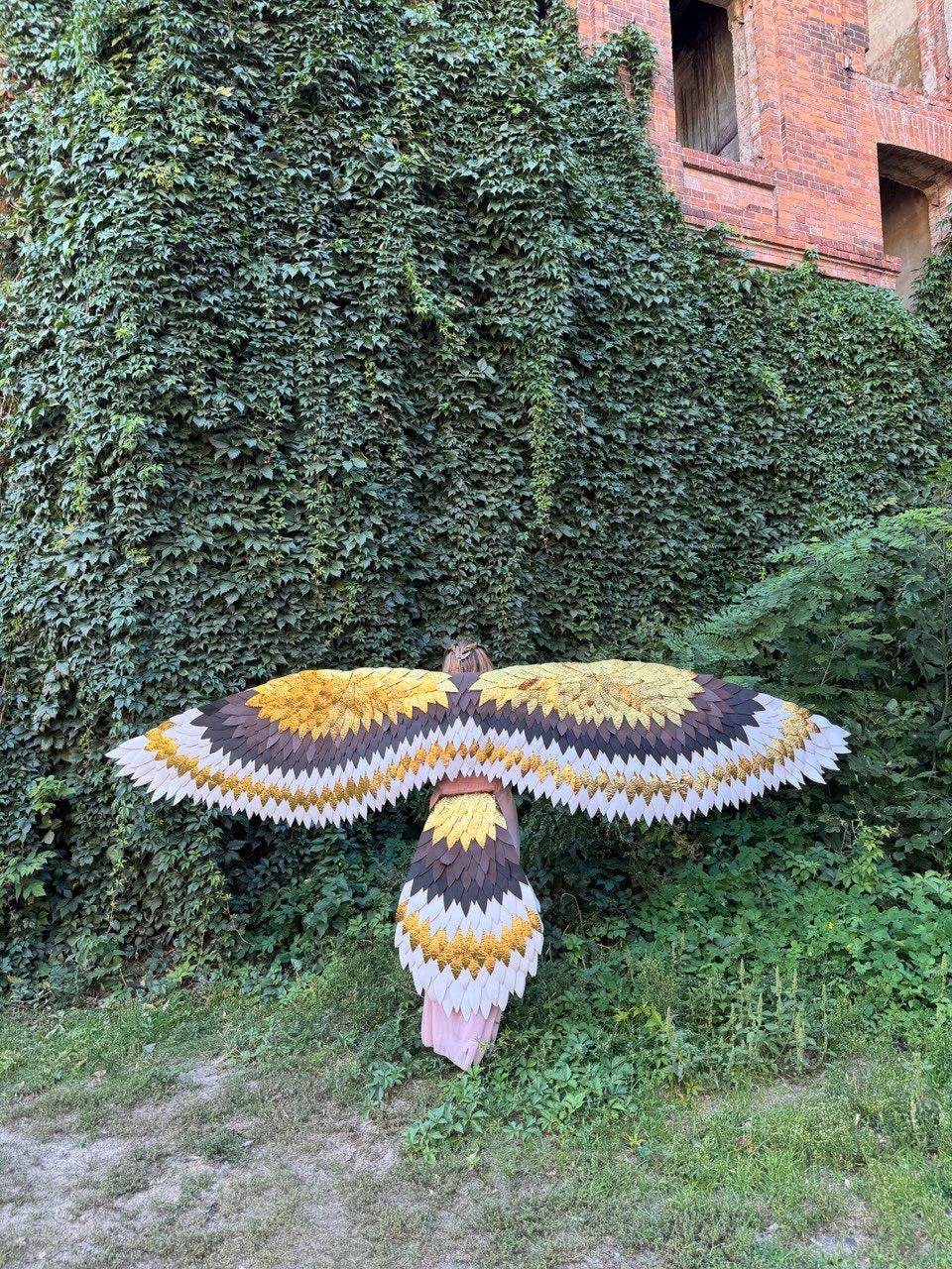 May include: A large, handcrafted bird sculpture with outstretched wings, featuring a layered design in shades of yellow, brown, and white. The sculpture is set against a backdrop of lush green foliage and a brick building.