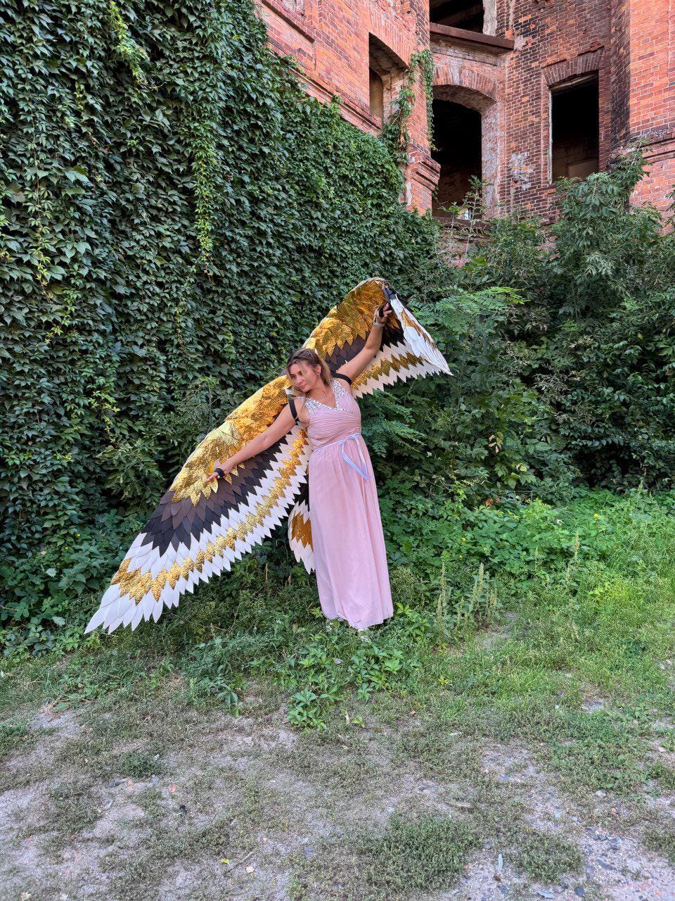 May include: A person wearing large, decorative wings with gold, white, and black segments. The wings are spread wide against a backdrop of green foliage and a brick building. The person is wearing a pink dress.