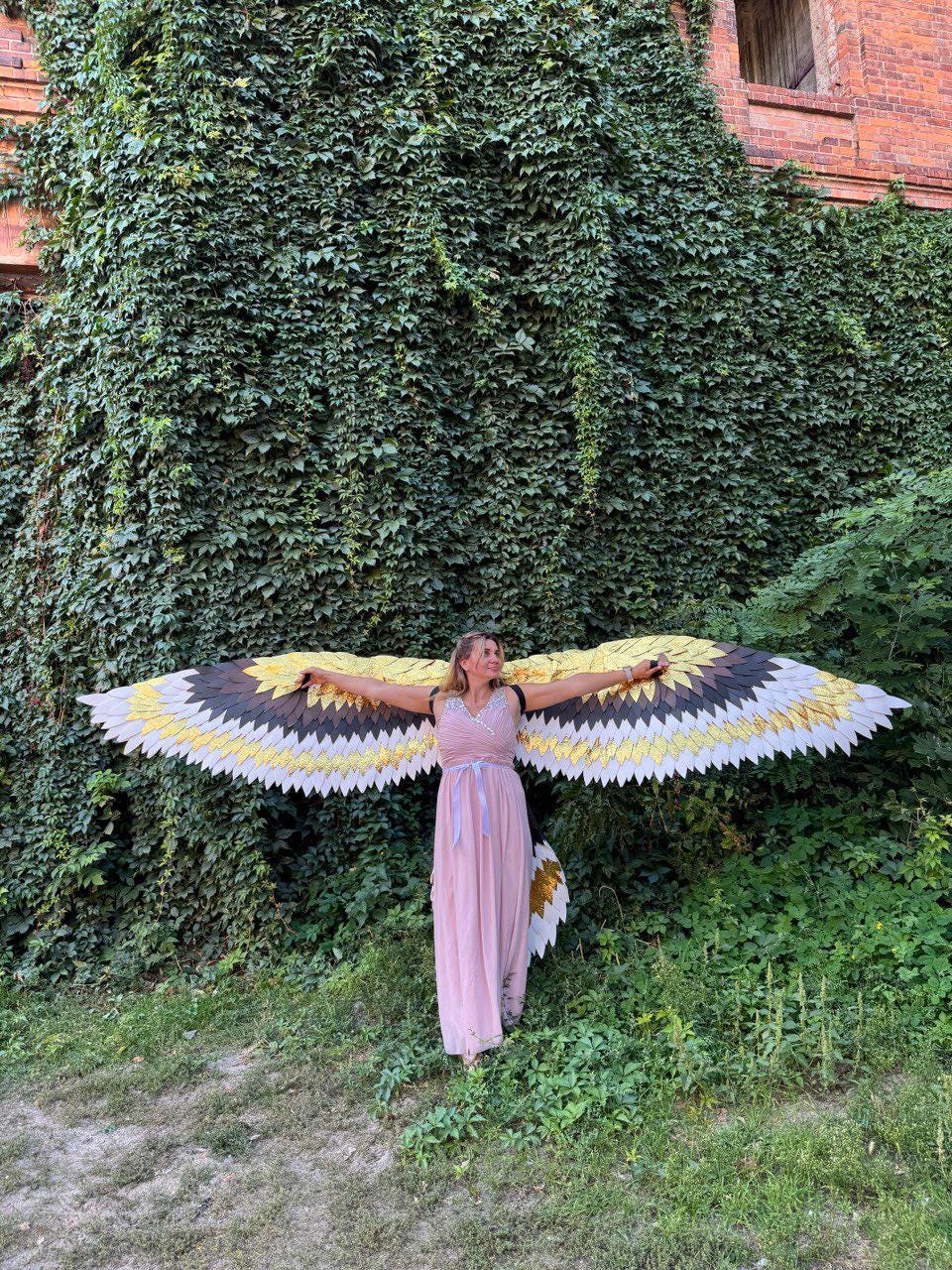 May include: A person wearing large, decorative bird wings with a black, gold, and white feather design. The wings are spread wide, and the person is wearing a pink dress. The background features a wall covered in green ivy.