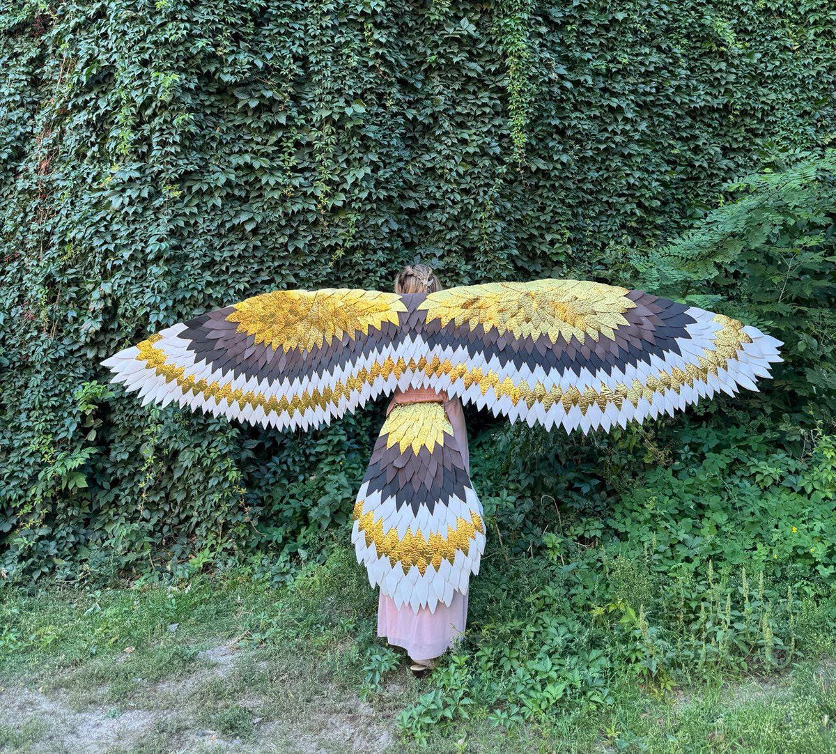 May include: A person wearing a large, decorative wing costume. The wings feature a layered design with white, brown, and gold-colored elements. The costume is set against a backdrop of green foliage.
