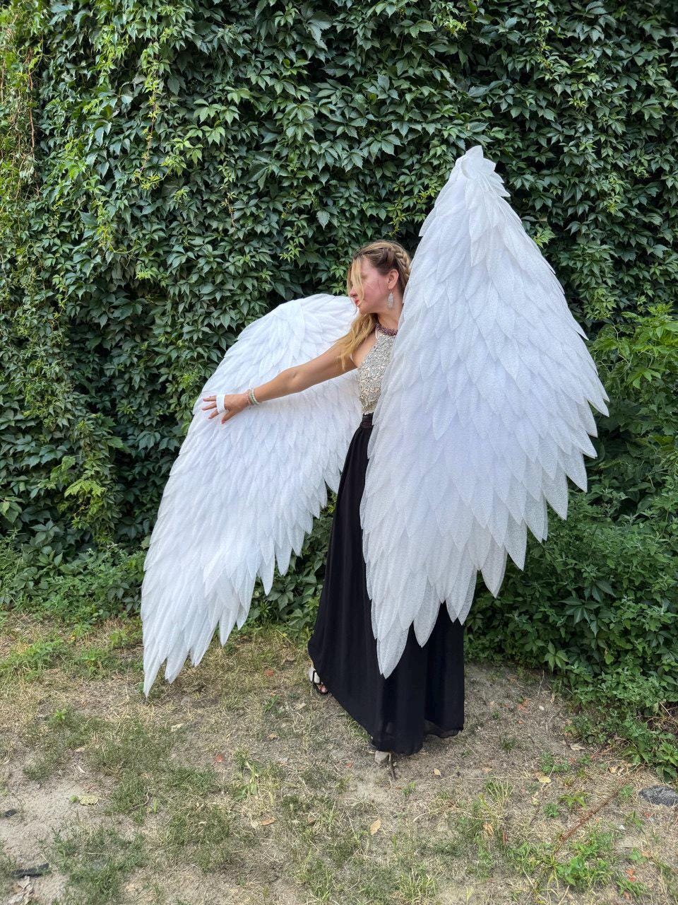 May include: Large, white angel wings with a feathered design. The wings are spread wide, showcasing their size and detail. The wings are the focal point, set against a backdrop of lush green foliage.