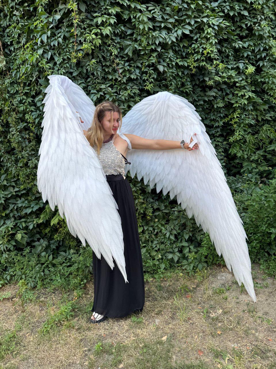 May include: A person wearing large, white feathered angel wings, standing in front of a green leafy background. The wings are spread wide, showcasing their size and detail. The person is wearing a black skirt and a white top.