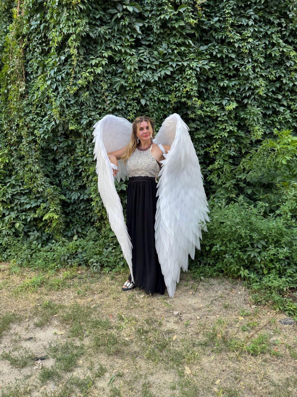 May include: A person wearing large, white angel wings, a silver top, and a black skirt, standing in front of a wall of green foliage. The wings are detailed with individual feathers.