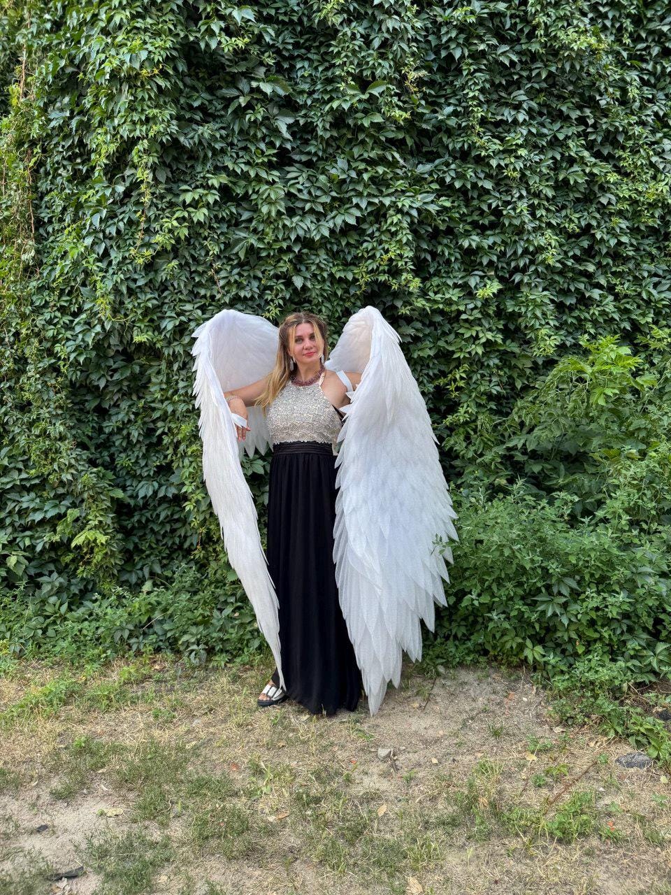 May include: A person wearing large, white angel wings, a silver and white top, and a long black skirt. The wings are detailed with individual feathers. The background is a lush green wall of foliage.